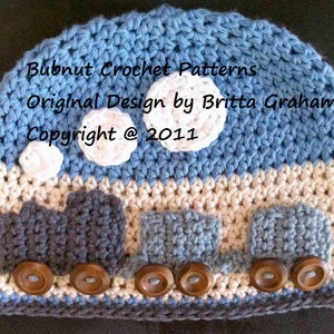 May include: A blue and white crocheted baby hat with a train design. The hat features three train carriages with brown buttons for wheels. The hat also has white crocheted clouds on the top. The text "Bubnut Crochet Patterns Original Design by Britta Graham Copyright @ 2011" is visible on the hat.