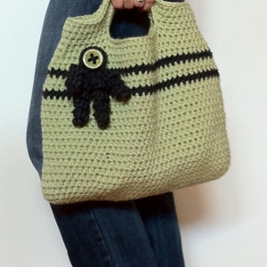 Puede incluir: Un bolso de mano de crochet con rayas negras y una flor de tela negra con un bot&oacute;n en el centro. El bolso lo lleva una persona que lleva pantalones vaqueros azules.