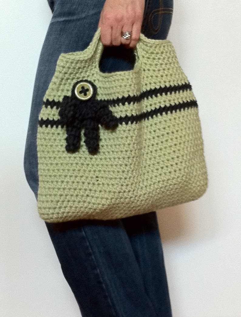 Puede incluir: Un bolso de mano de crochet con rayas negras y una flor de tela negra con un bot&oacute;n en el centro. El bolso lo lleva una persona que lleva pantalones vaqueros azules.