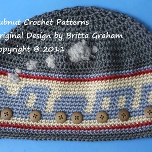 Könnte beinhalten: Graue Häkelmütze mit einem blau-weißen Streifen und einer Reihe von Holzknöpfen. Die Mütze trägt den Text "Bubnut Crochet Patterns Original Design by Britta Graham Copyright @ 2011" auf der Oberseite.