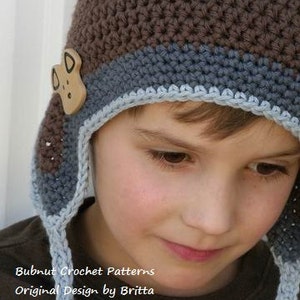 May include: A brown and blue crocheted hat with a small brown animal applique. The hat has a long tie that hangs down the front of the hat. The hat is worn by a child.