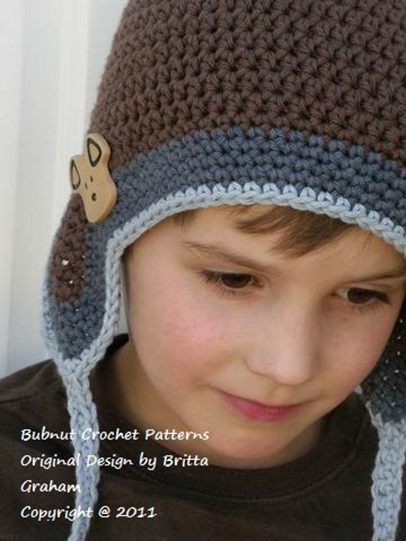 May include: A brown and blue crocheted hat with a small brown animal applique. The hat has a long tie that hangs down the front of the hat. The hat is worn by a child.