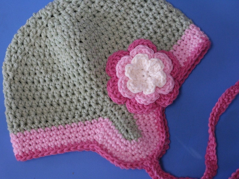 May include: A crocheted hat with a pink flower and a pink and green trim. The hat has a long pink tie.