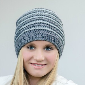 May include: A woman wearing a gray and blue striped crocheted beanie hat. The hat has a simple design with alternating stripes of gray and light blue yarn.
