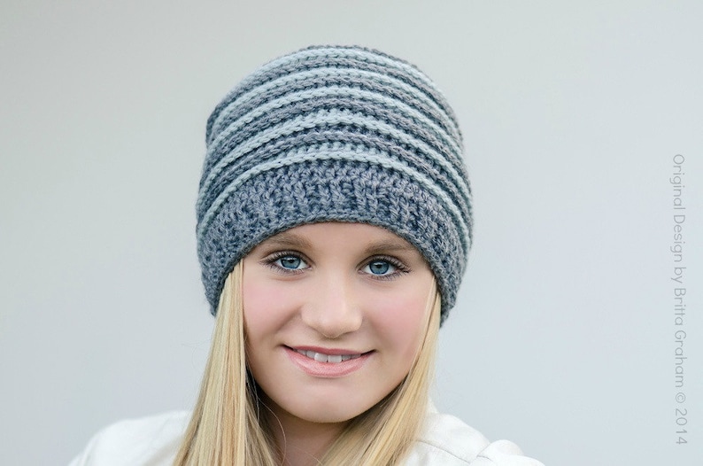 May include: A woman wearing a gray and blue striped crocheted beanie hat. The hat has a simple design with alternating stripes of gray and light blue yarn.