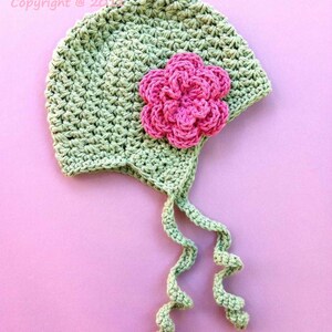 Op de afbeelding: Een groene gehaakte baby muts met een roze gehaakte bloem. De muts heeft een lange strik die aan de achterkant is geknoopt. De muts ligt op een roze achtergrond. Bubnut Crochet Patterns Original Design by Britta Graham Copyright @ 2014