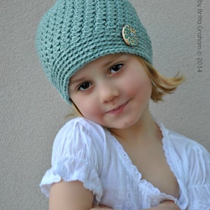 May include: A young person wearing a light blue crocheted hat with a floral button. The hat has a slouchy style and is pulled down over the forehead. The person is wearing a white shirt with lace trim.