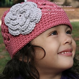 Puede incluir: Una niña pequeña lleva un gorro de punto rosa con una flor de punto gris en la parte superior. El gorro tiene un diseño sencillo y está hecho con un hilo suave. La niña sonríe y mira hacia arriba.
