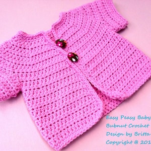 May include: A pink crocheted baby cardigan with short sleeves and two buttons. The cardigan is on a pink background. Easy Peasy Baby Cardi Bubnut Crochet Patterns Design by Britta Graham Copyright @ 2012