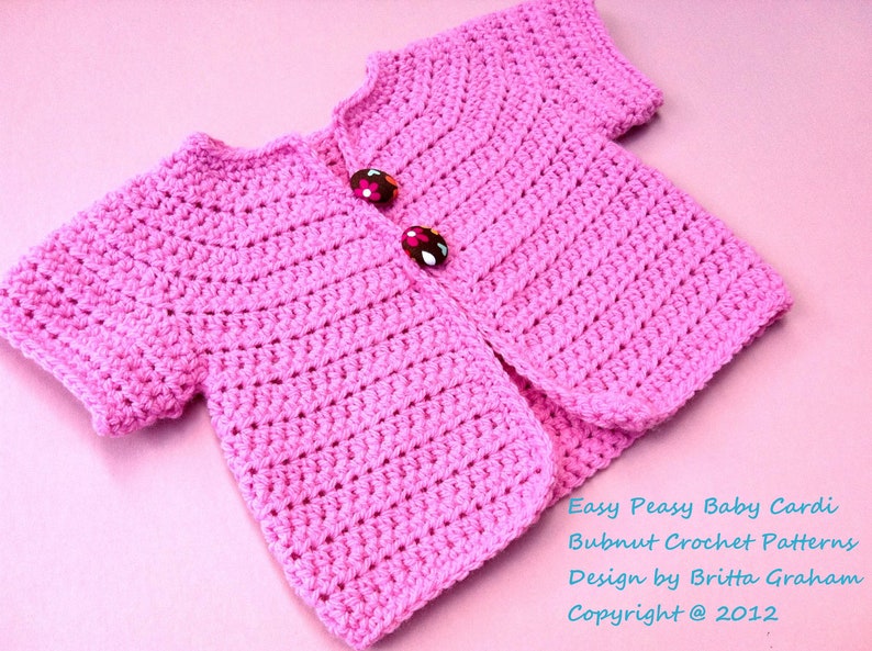 May include: A pink crocheted baby cardigan with short sleeves and two buttons. The cardigan is on a pink background. Easy Peasy Baby Cardi Bubnut Crochet Patterns Design by Britta Graham Copyright @ 2012