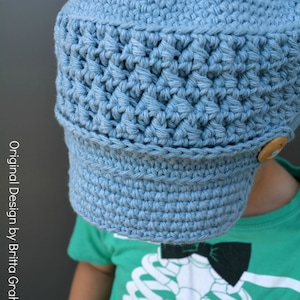 May include: A light blue crocheted hat with a brim and a button on the side. The hat is worn by a child wearing a green shirt with a skeleton graphic.