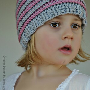 May include: A young girl wearing a gray and pink striped knitted hat. The hat has a wide brim and covers her ears. She is wearing a white shirt with lace trim.