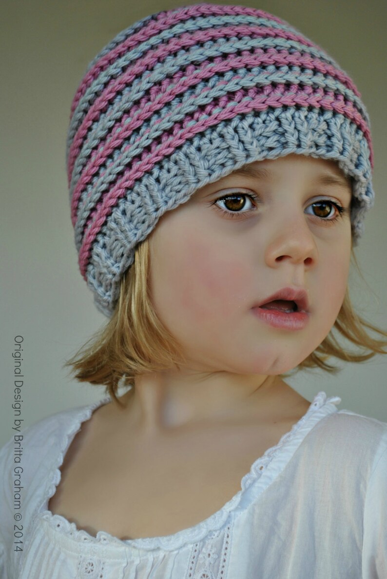 May include: A young girl wearing a gray and pink striped knitted hat. The hat has a wide brim and covers her ears. She is wearing a white shirt with lace trim.