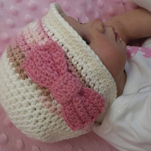 Puede incluir: Un gorro de bebé de ganchillo blanco con una banda de rayas rosa y marrón y un gran lazo de ganchillo rosa.