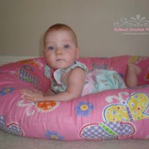 Könnte beinhalten: Ein rosa-weißes Babykissen mit Schmetterlingsmuster. Das Kissen ist mit Füllmaterial gefüllt und hat eine weiche, komfortable Textur. Es ist ideal für Babys, die das Sitzen oder Rollen lernen.