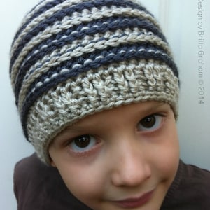 May include: A young person wearing a striped crocheted hat in shades of gray and beige. The hat is pulled down low over their forehead, and they are looking at the camera with a slight smile.