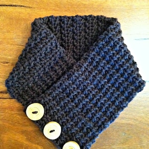 May include: A dark gray knitted cowl neck scarf with three wooden buttons.  Original Design by Britta Graham © 2011.