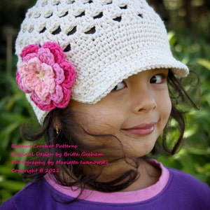 Puede incluir: Una niña pequeña con un sombrero blanco de ganchillo con una flor rosa en un lado. El sombrero tiene un ala ancha y un diseño de ganchillo. La niña sonríe y mira hacia un lado. El sombrero es un diseño de patrón de ganchillo de Britta Graham. Fotografía de Marietta Iwanowski. Copyright 2011.