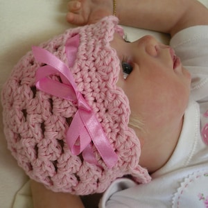 Puede incluir: Un gorro de bebé de ganchillo rosa con un lazo de satén rosa. El gorro lo lleva un bebé con el pelo rubio.