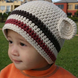 May include: A beige crocheted hat with brown and burgundy stripes and two ear flaps. The hat is worn by a child.