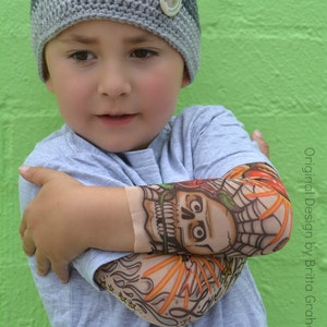 May include: A young person wearing a gray and white crocheted hat with a monkey button on the side. The person is wearing a light blue t-shirt with a colorful skull and flames tattoo sleeve.