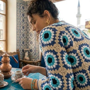 Könnte beinhalten: Eine Frau trägt eine beige, blau und weiße Häkeljacke mit Blumenmuster. Sie sitzt an einem blau gefliesten Tisch und hält eine kleine Tasse und Untertasse. Goldene Ohrringe und Armbänder sind sichtbar.
