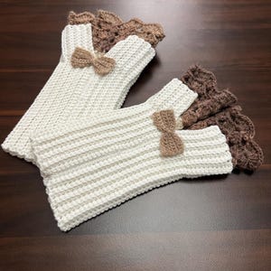 May include: Handmade crochet fingerless gloves in cream and brown. The cream-colored gloves have a ribbed texture and are adorned with small brown bows. The cuffs are a darker brown with a scalloped edge. The gloves are displayed on a dark wood surface.