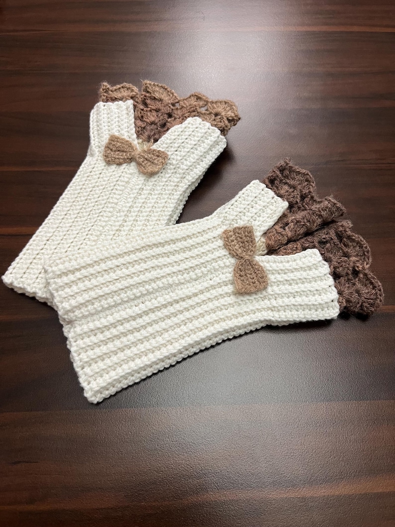 May include: Handmade crochet fingerless gloves in cream and brown. The cream-colored gloves have a ribbed texture and are adorned with small brown bows. The cuffs are a darker brown with a scalloped edge. The gloves are displayed on a dark wood surface.