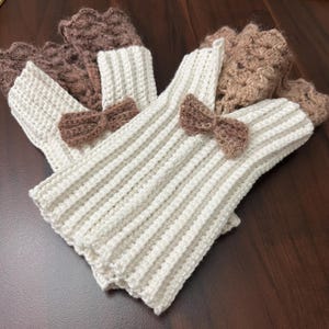 May include: A set of ivory-colored, ribbed knit leg warmers with brown crochet bow accents. The leg warmers have a brown crochet cuff at the top. The leg warmers are displayed on a dark brown surface.