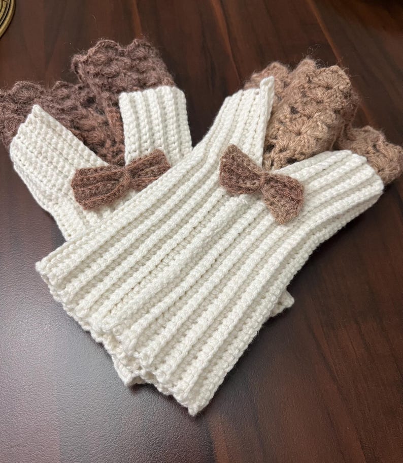 May include: A set of ivory-colored, ribbed knit leg warmers with brown crochet bow accents. The leg warmers have a brown crochet cuff at the top. The leg warmers are displayed on a dark brown surface.