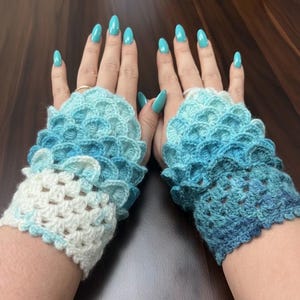 Könnte beinhalten: Ein Paar handgefertigte, gehäkelte Fingerhandschuhe in Blau- und Weißtönen. Die Handschuhe haben ein wellenförmiges Design und werden auf einer dunklen Holzoberfläche präsentiert. Der Farbverlauf geht von Weiß über Hellblau zu Dunkelblau.