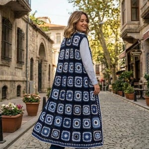 May include: A long, navy blue crochet vest with a geometric pattern of squares and circles. The vest is worn over a white shirt and jeans. The crochet design features shades of blue and white.