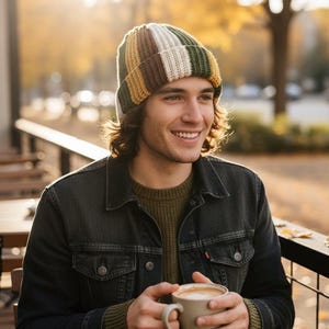 以下が含まれることがあります： 緑、茶色、白、黄色のストライプ柄のビーニー。濃い色のデニムジャケットを着て、ベージュのマグカップに入ったコーヒーを持っています。背景は秋の色でぼやけています。