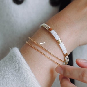 Puede incluir: Primer plano de una muñeca con dos pulseras de cadena dorada y una pulsera de cuentas blancas y doradas.