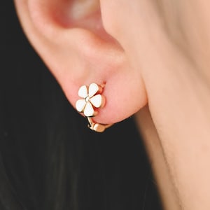 May include: A pair of gold hoop earrings with a white flower design. The earrings are small and delicate.