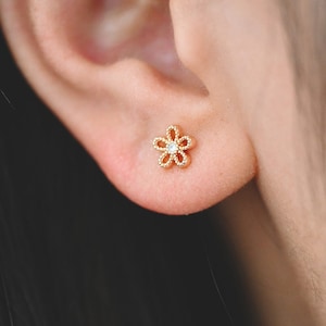 May include: A close-up of a gold-colored flower-shaped stud earring with a small, clear gemstone in the center. The earring is worn on an ear, with the person's dark hair visible in the background. The earring has a delicate, openwork design.