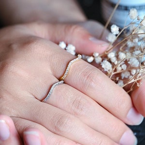May include: Two delicate, wavy rings with small, clear stones set in gold and silver. The rings are worn on the finger of a hand resting on a black fabric background.