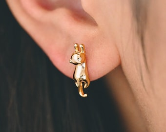 May include: A gold-colored cat-shaped earring is shown in the image. The earring is a stud, and the cat is depicted in a standing pose. The earring is worn on an ear, and the background is blurred.