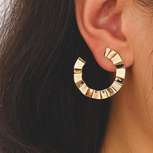 Peut inclure: Gros plan sur une boucle d'oreille dorée en forme de cercle avec un motif texturé et nervuré. La boucle d'oreille est en forme de demi-cercle et est portée sur l'oreille. L'arrière-plan est de couleur claire et neutre.