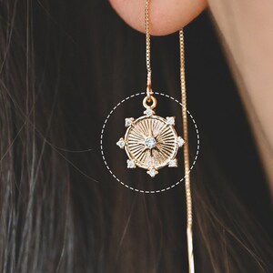 May include: A gold-colored threader earring with a circular pendant. The pendant features a sunburst design with a central clear gemstone and smaller gemstones around the edge. The earring is shown worn in an ear.