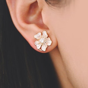 May include: A pair of gold flower-shaped earrings with white enamel and a clear rhinestone in the centre.