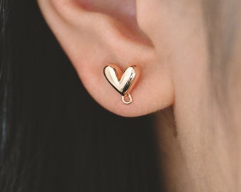 May include: Gold heart-shaped earrings with a small dangling charm. The earrings are worn in a person's ear.
