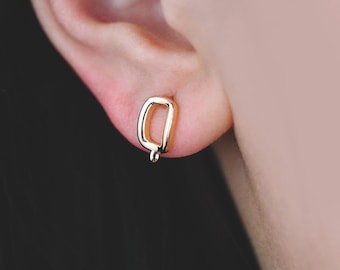 Puede incluir: Primer plano de un pendiente dorado con forma de la letra «Q» en una oreja. El pendiente es un pequeño botón y la oreja está sobre un fondo borroso. Joyería.
