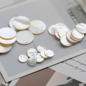 May include: A collection of iridescent, round mother-of-pearl shell beads. The beads vary in size and are arranged in small piles on a light gray surface, with a few lying on a printed paper. Each bead has a small hole.