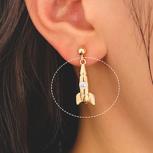 May include: Gold rocket ship dangle earrings with a clear rhinestone. The earrings are hanging from a person's ear.