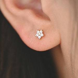May include: A close-up of a person's ear with a small gold flower stud earring with clear cubic zirconia stones.