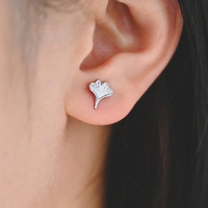 May include: A pair of silver ginkgo leaf stud earrings. The earrings are small and delicate, and they feature a detailed design of a ginkgo leaf.