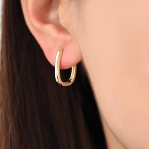 Puede incluir: Primer plano de un pendiente de aro rectangular dorado que se lleva en una oreja. El pendiente tiene un acabado pulido y un diseño sencillo y moderno. El fondo está borroso, centrando la atención en la joya.