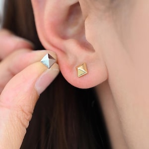 Puede incluir: Un par de pequeños pendientes de forma piramidal geométrica. Los pendientes están hechos de metal dorado y se llevan en los lóbulos de las orejas de una persona.