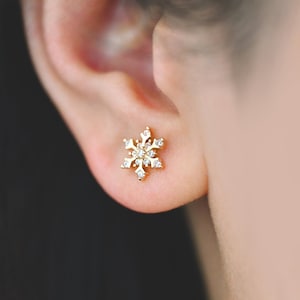 May include: A close-up of a gold-colored snowflake stud earring with small, clear gemstones. The earring is worn on an ear, with the person's hair and skin visible in the background. The earring is a winter-themed accessory.